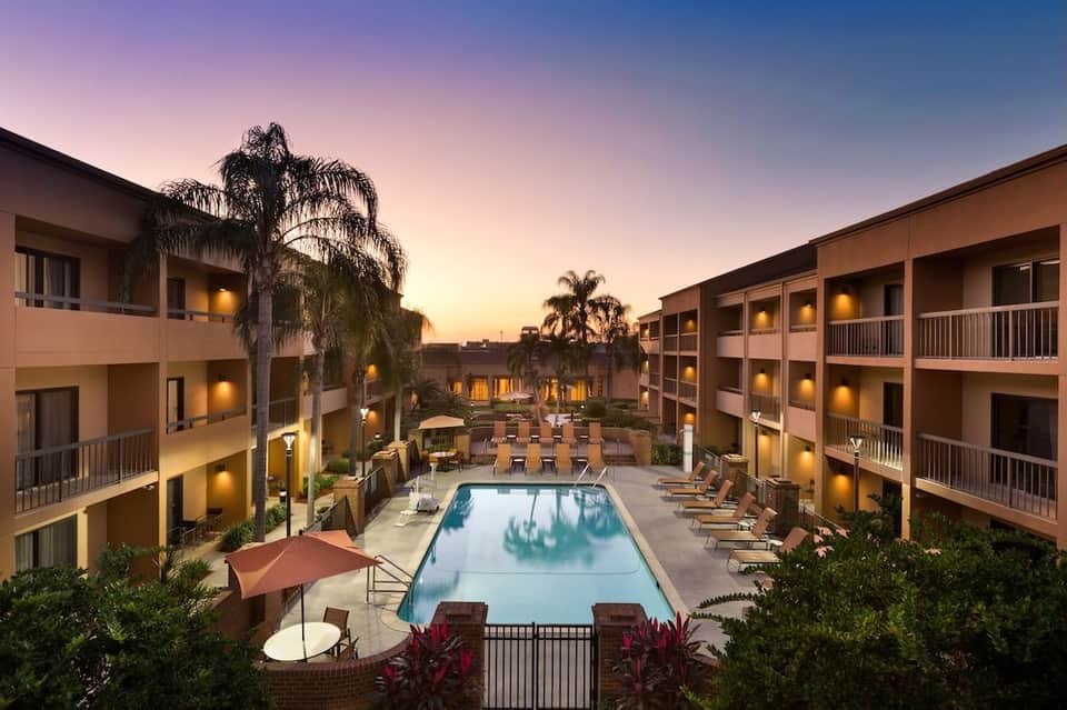 Resort pool at dusk with illuminated building wings, palm trees, and lounge chairs under purple twilight sky
