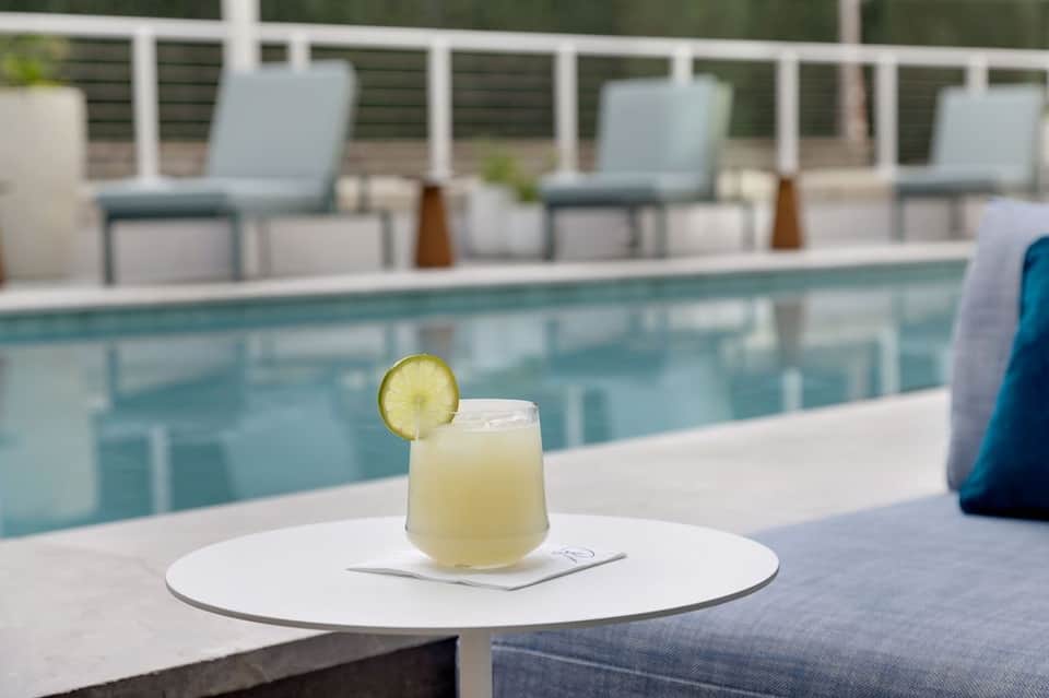 Lime-garnished cocktail on white table by pool with loungers and hedges in background
