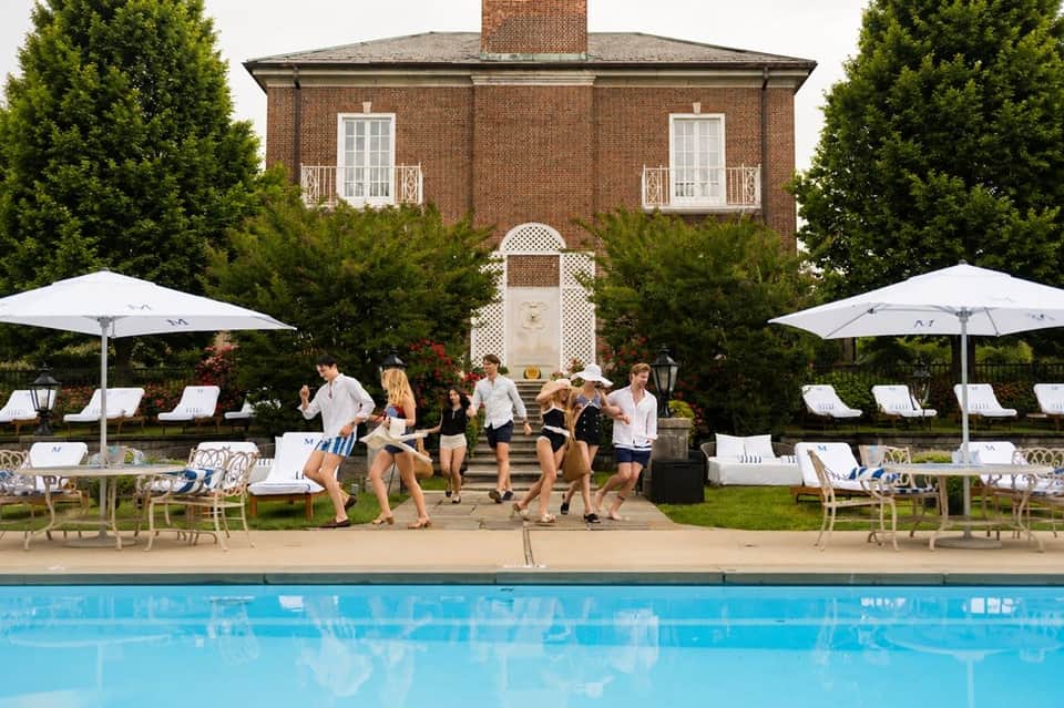 Resort pool area with guests playing cornhole, white umbrellas, lounge chairs, and brick mansion in background