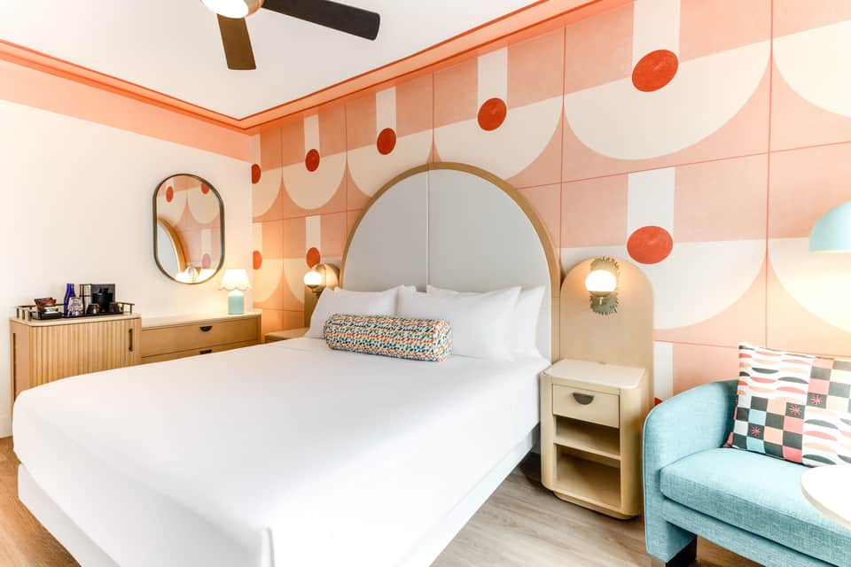 Modern hotel bedroom with coral accent wall, arched headboard, and turquoise seating