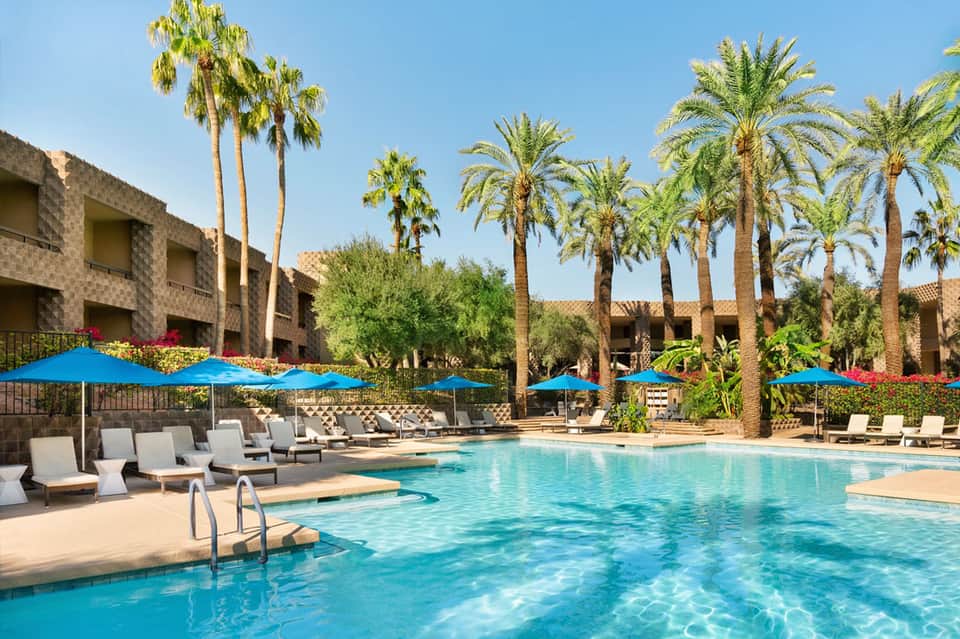 Resort pool with crystal blue water, palm trees, lounge chairs, blue umbrellas, and tan stone buildings under clear sky