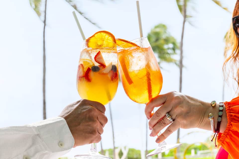 Two tropical cocktails with fruit garnish being toasted with palm trees background
