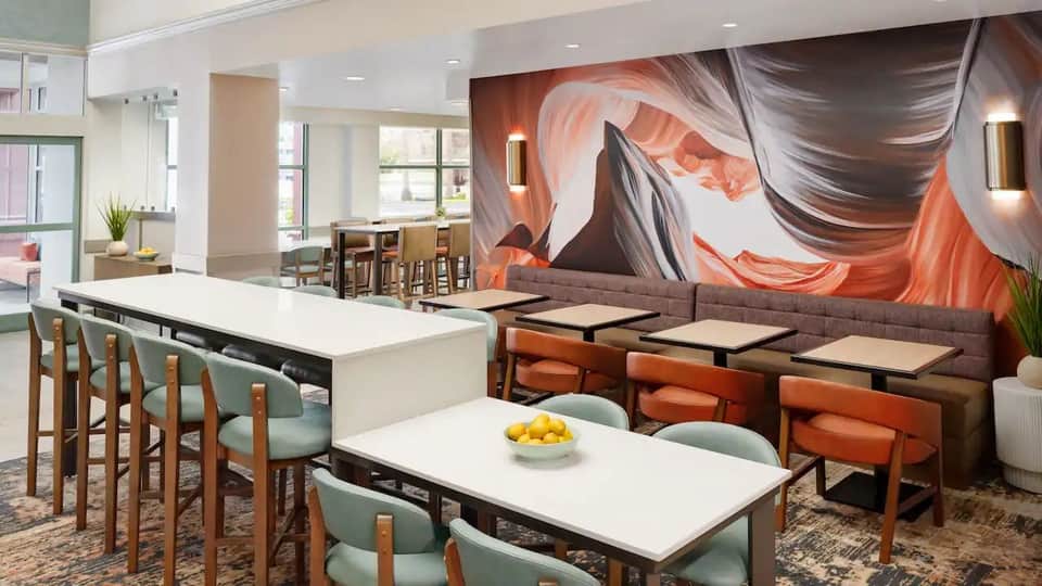Hotel lobby lounge with white dining tables, wooden chairs, large abstract orange wall art, and floor-to-ceiling windows