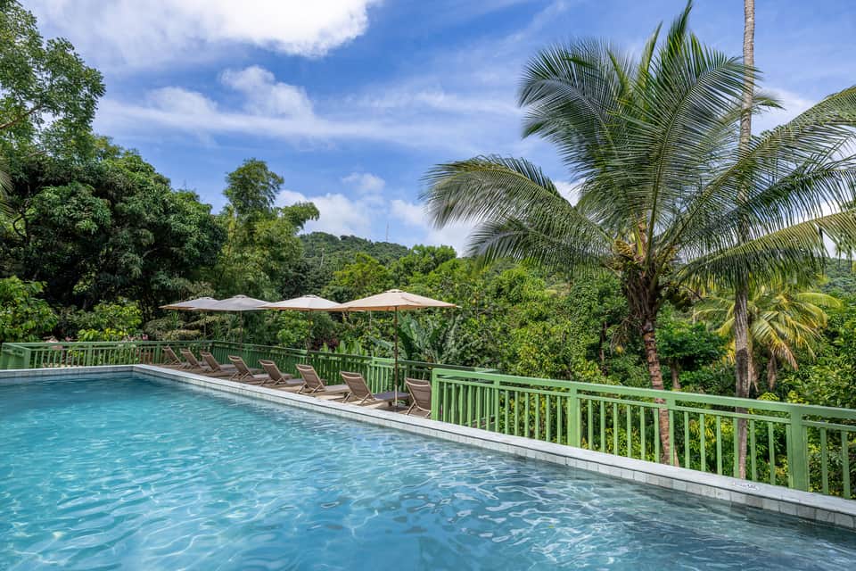Infinity pool with lounge chairs and umbrellas overlooking tropical mountains and palm trees under blue sky