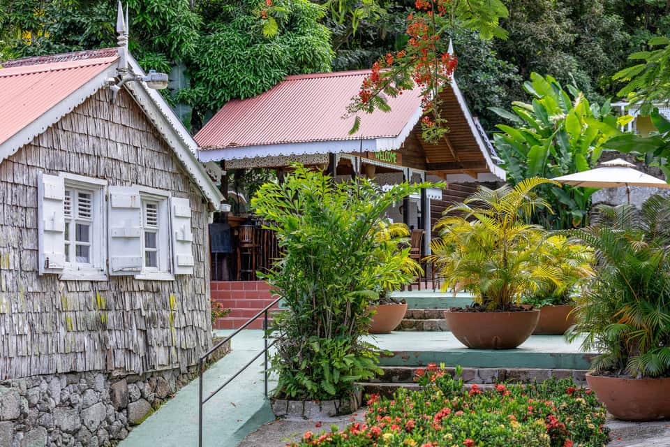 Colonial-style buildings with red roofs, wooden architecture, tropical plants, and stone pathway in garden setting