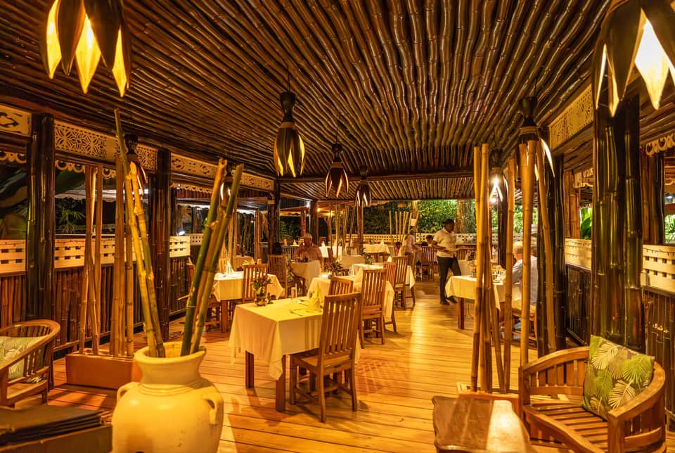 Warm-lit wooden dining pavilion with high ceilings, decorative bamboo, and formally set tables