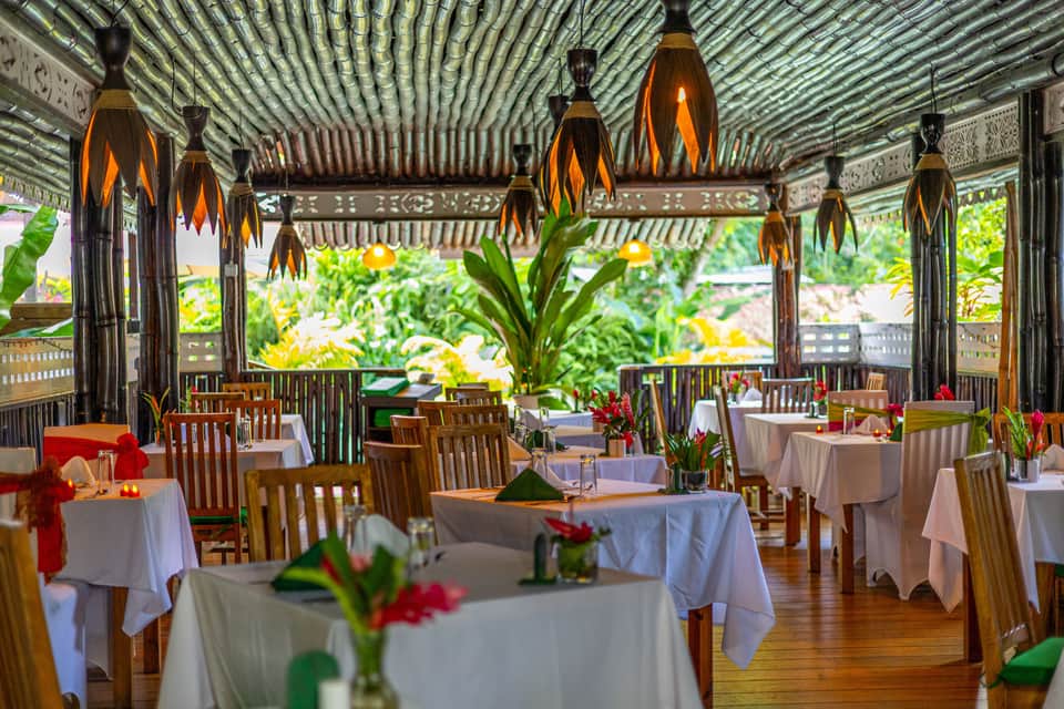 Fine dining room with white tablecloths, hanging bronze pendant lights, and tropical flower arrangements
