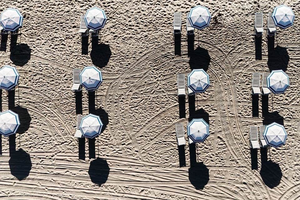 Aerial view of organized blue and white beach umbrellas with dark shadows on sand