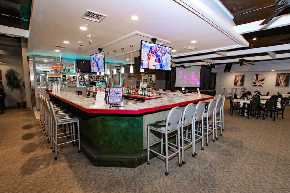Modern bar lounge with curved counter, red and green accents, white bar seating, flat-screen TV, and ambient lighting
