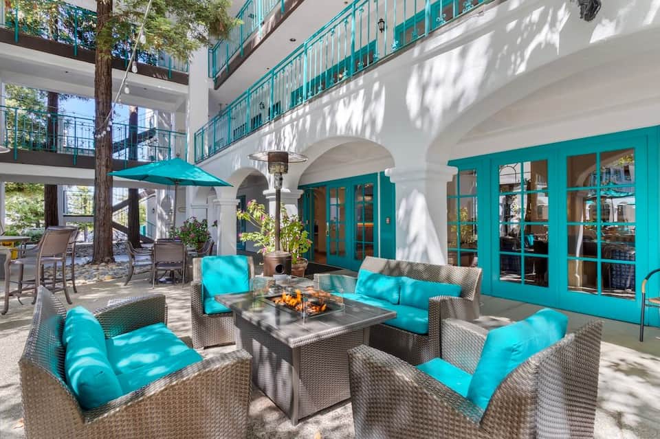Outdoor courtyard with turquoise lounge seating, fire pit table, tall tree, white arches, and turquoise doors