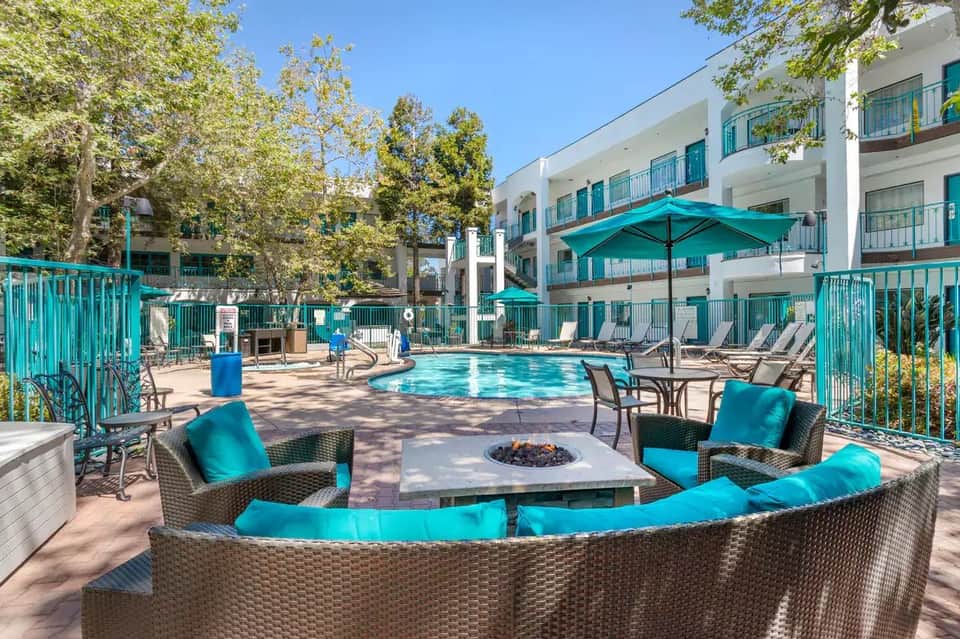Pool area with fire pit seating, turquoise cushioned furniture, white buildings, and mature shade trees