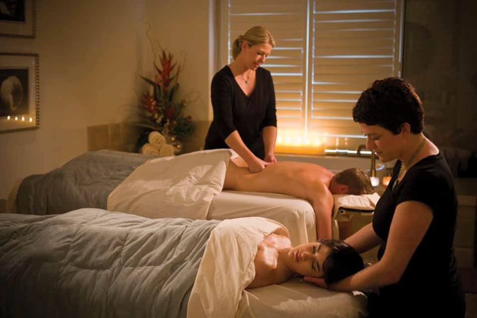 Two massage therapists performing treatments on clients lying on massage beds in a spa room with warm lighting