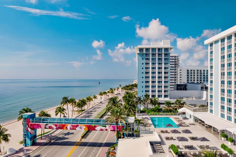 Beachfront hotel resort with pool, palm trees, colorful public art installation, and ocean view