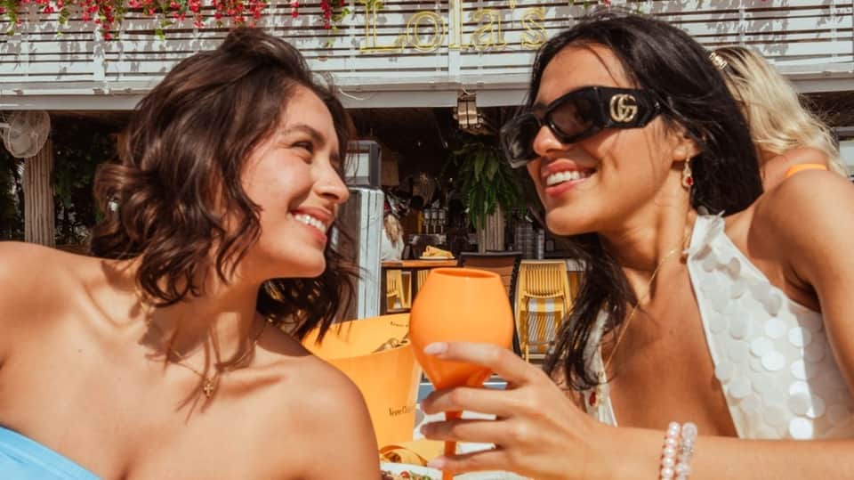 Two smiling women in bikinis holding tropical drinks at upscale beach club bar