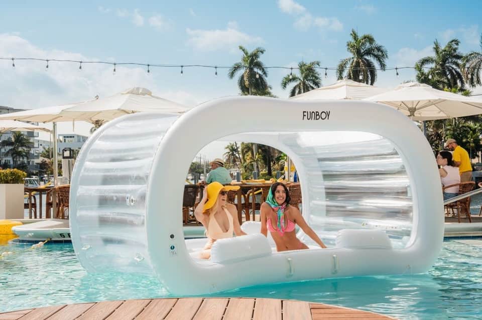 Transparent bubble lounger in resort pool with two people inside, palm trees and white umbrellas in background