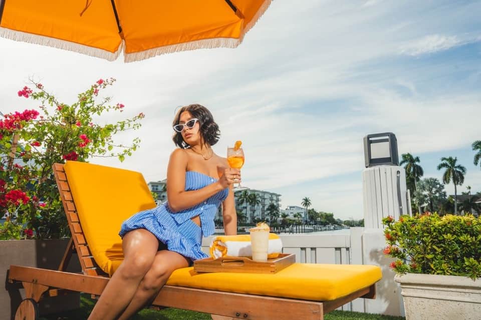 Woman in blue striped dress sitting under yellow umbrella holding tropical drink by waterfront