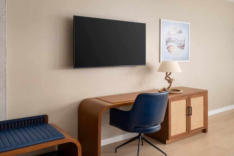 Hotel room workspace with mounted TV, wooden desk, navy chair, and nautical-themed wall art