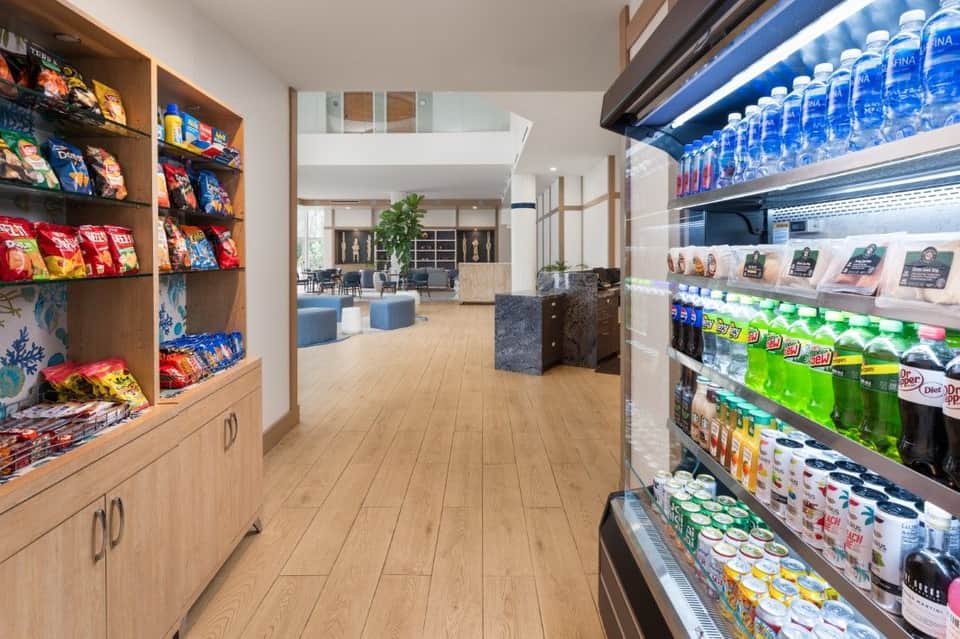 Modern hotel convenience store with snack shelves on left, refrigerated beverages on right, and lounge area visible in background