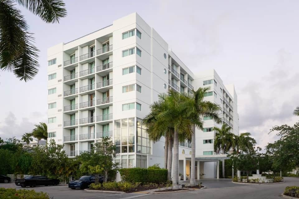 White mid-rise residential building with balconies, palm trees, and covered carport entrance