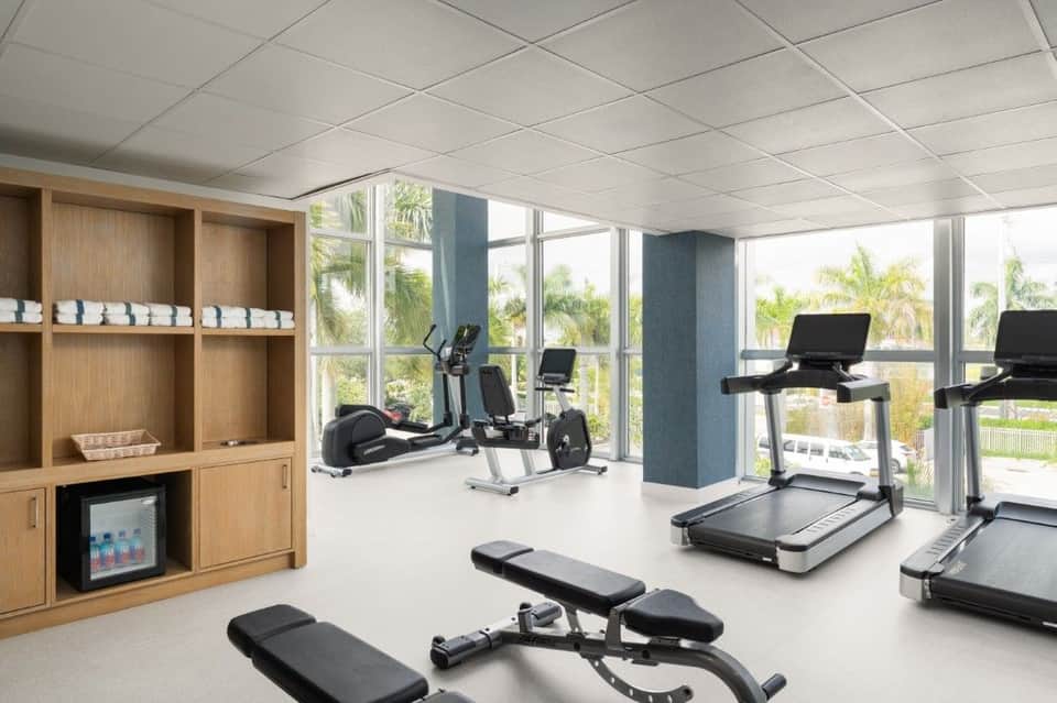 Bright fitness center with cardio equipment, large windows overlooking palm trees, and wooden storage cabinets