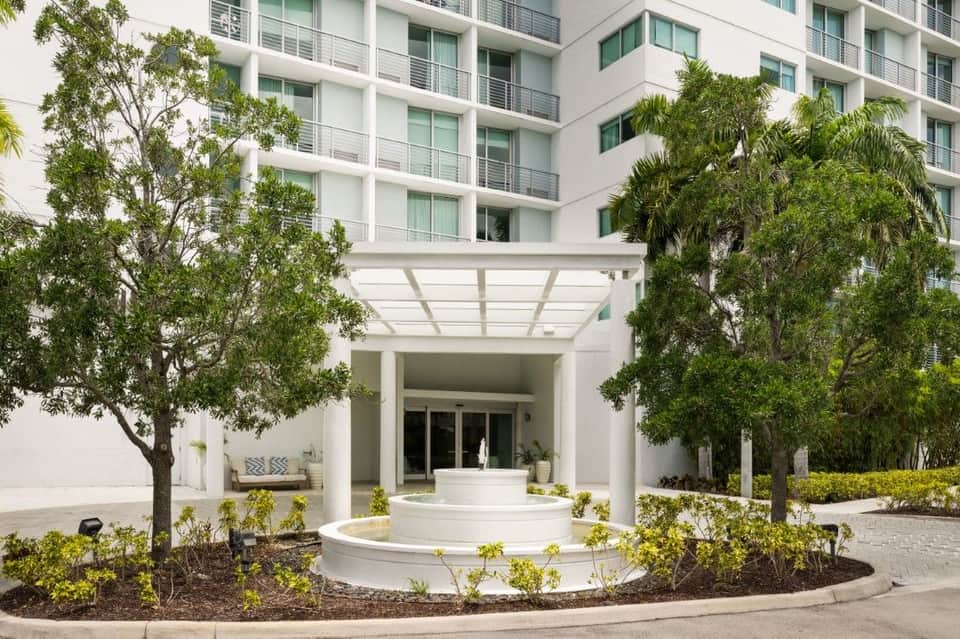 Modern hotel entrance with white columns, covered pergola, circular fountain, and mature landscaping