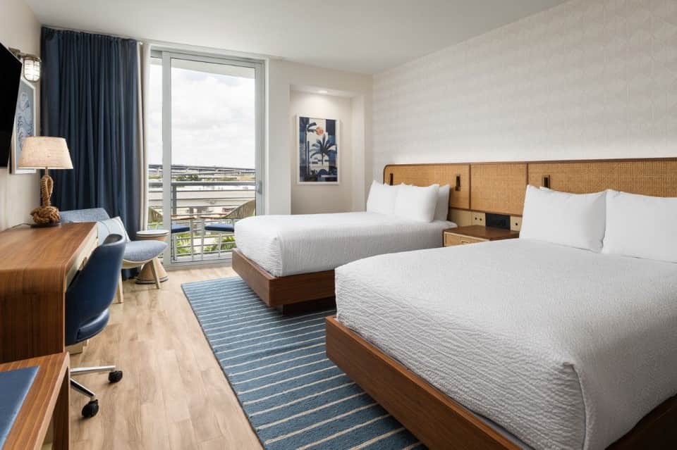 Spacious hotel room with two queen beds, woven headboards, balcony with bay views, and blue accents