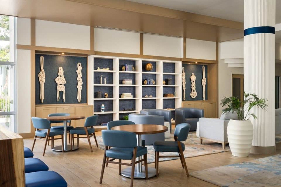 Modern hotel lounge with blue chairs, wooden tables, wall-mounted macramé art, built-in shelving, and natural light from floor-to-ceiling windows
