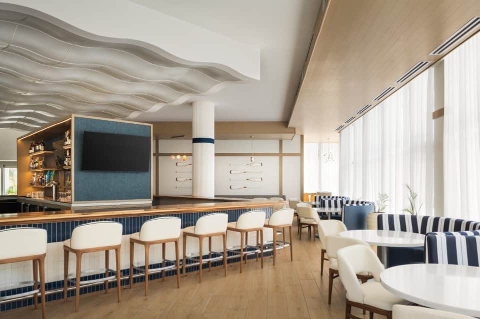 Contemporary hotel bar and lounge with wavy ceiling design, bar seating, striped upholstered chairs, and a TV mounted on blue accent wall