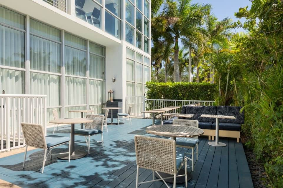 Outdoor terrace with woven lounge chairs, blue cushions, palm trees, and modern glass building facade with white railings