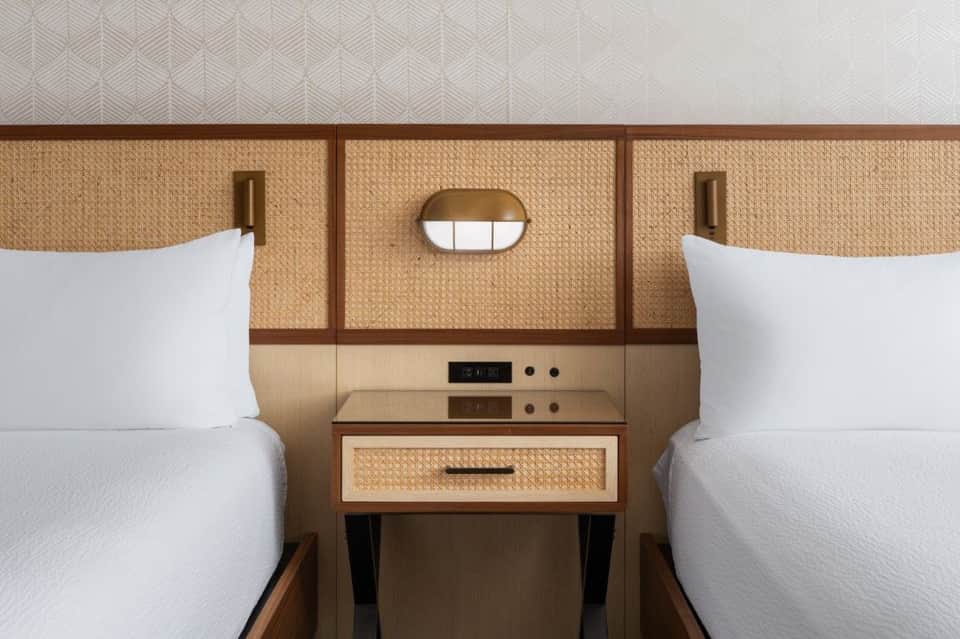 Twin hotel beds with woven rattan headboards separated by integrated wooden nightstand with drawer and round mirror