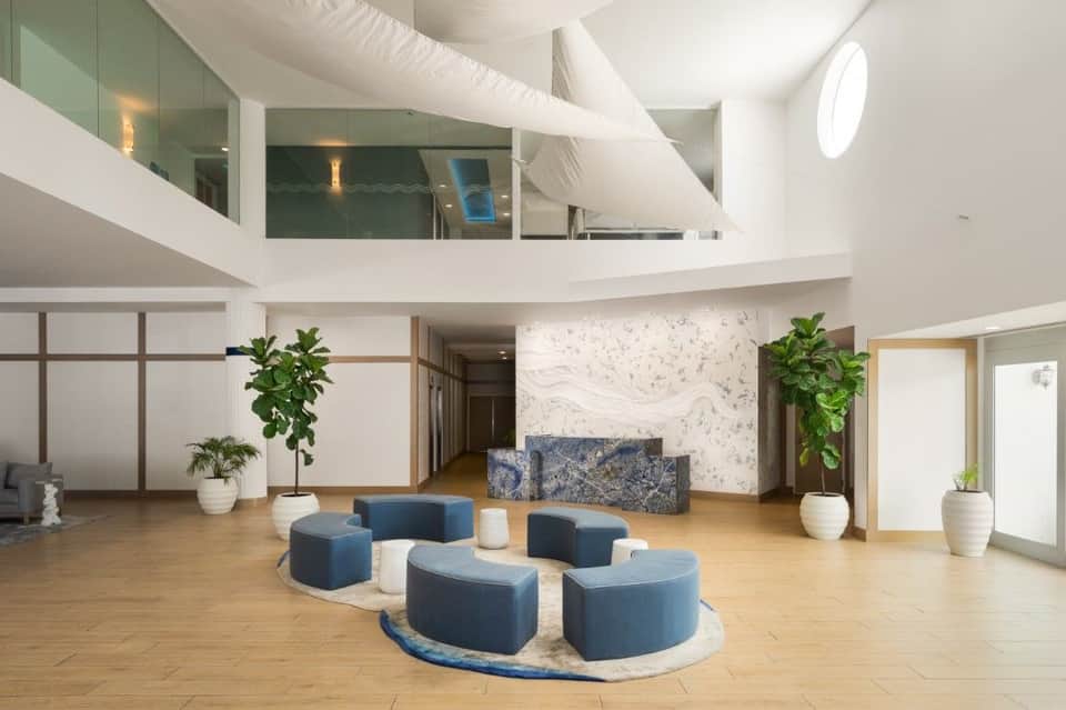 Multi-level hotel lobby atrium with curved white architectural elements, blue seating pods, marble accent wall, potted plants, and natural lighting