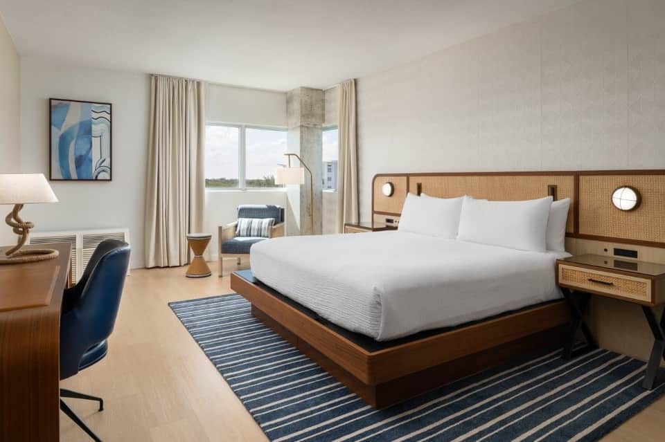 Modern hotel suite with rattan headboard bed, waterfront balcony view, white bedding, blue striped rug, desk workspace, and contemporary furnishings