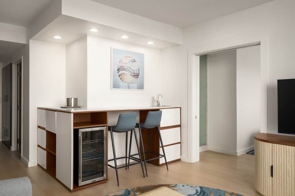 Hotel kitchenette with wooden cabinetry, white countertop, built-in wine cooler, bar seating, and blue-upholstered stools