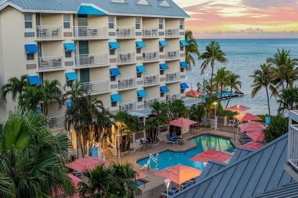 Multi-story beachfront hotel with blue awnings, pool, colorful umbrellas at sunset