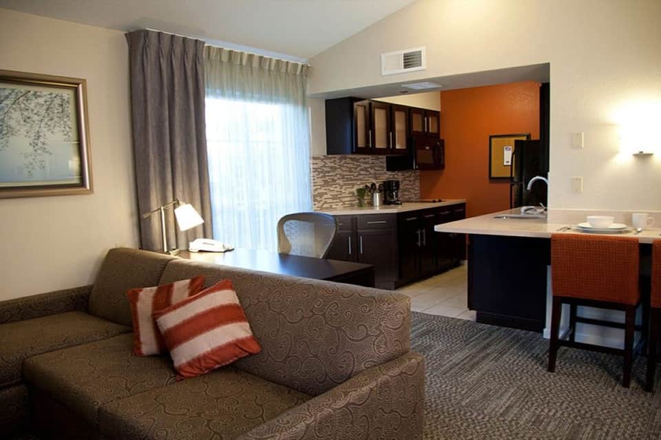 Hotel suite with living area featuring tan sofa, kitchenette with dark cabinets, orange accent wall, and large windows with curtains