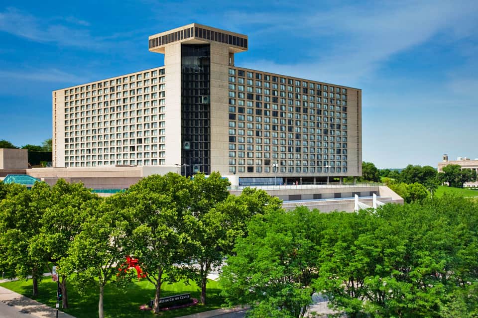 Modern brutalist hotel tower with distinctive vertical core rising above manicured gardens under blue sky
