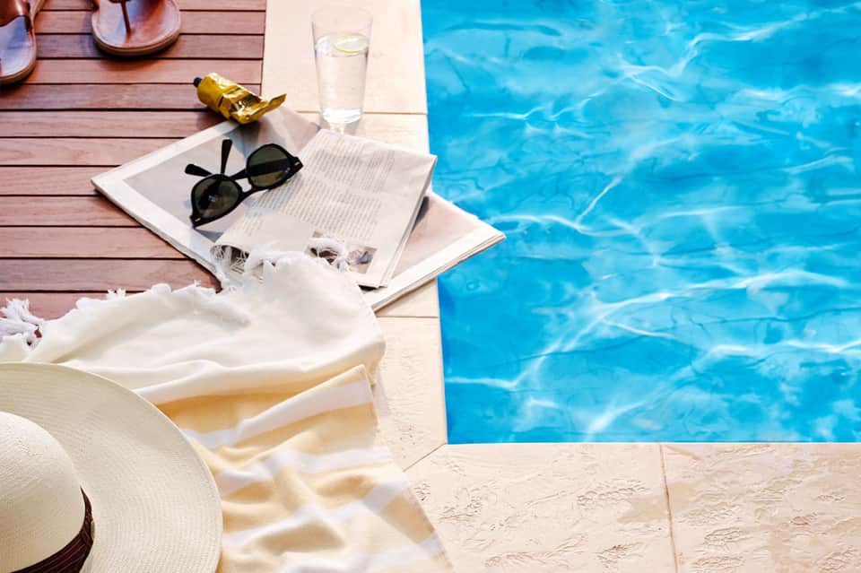 Poolside relaxation scene with sunglasses, newspaper, water glass, and white towel on wooden deck next to clear blue water