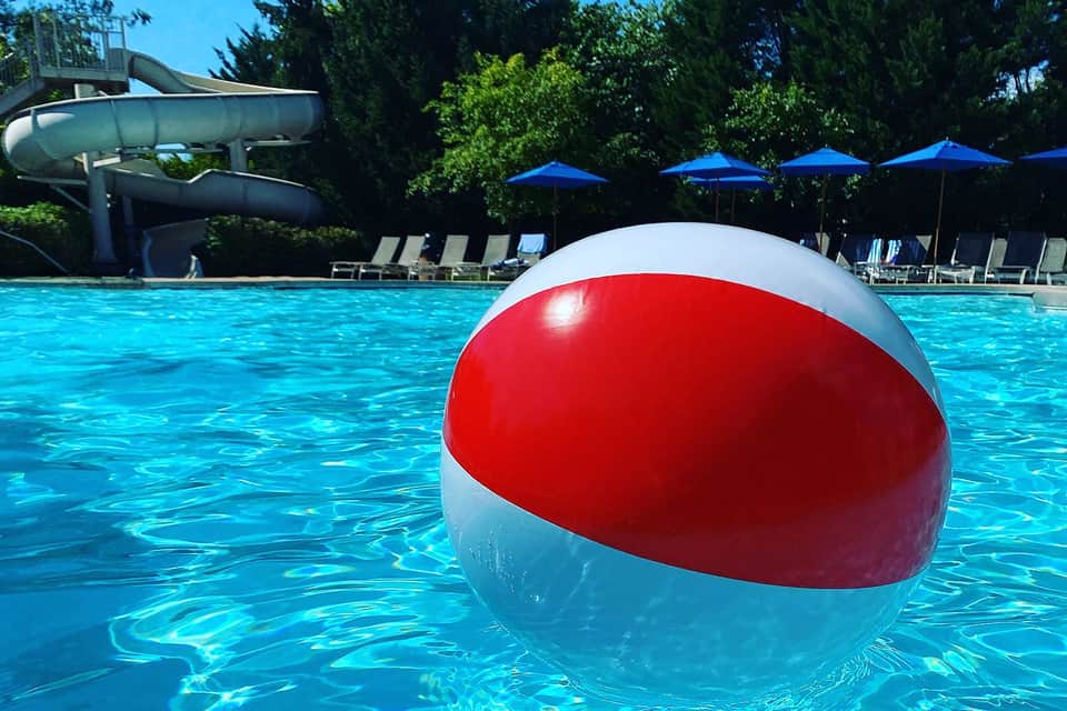 Red and white beach ball floating in a crystal-clear resort pool with waterslide in background