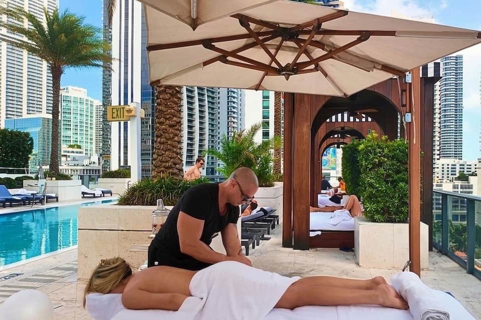Outdoor poolside massage service with therapist treating client under beige umbrella