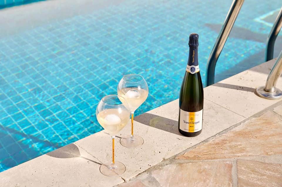 Two champagne glasses and bottle on pool deck edge with clear blue water in background