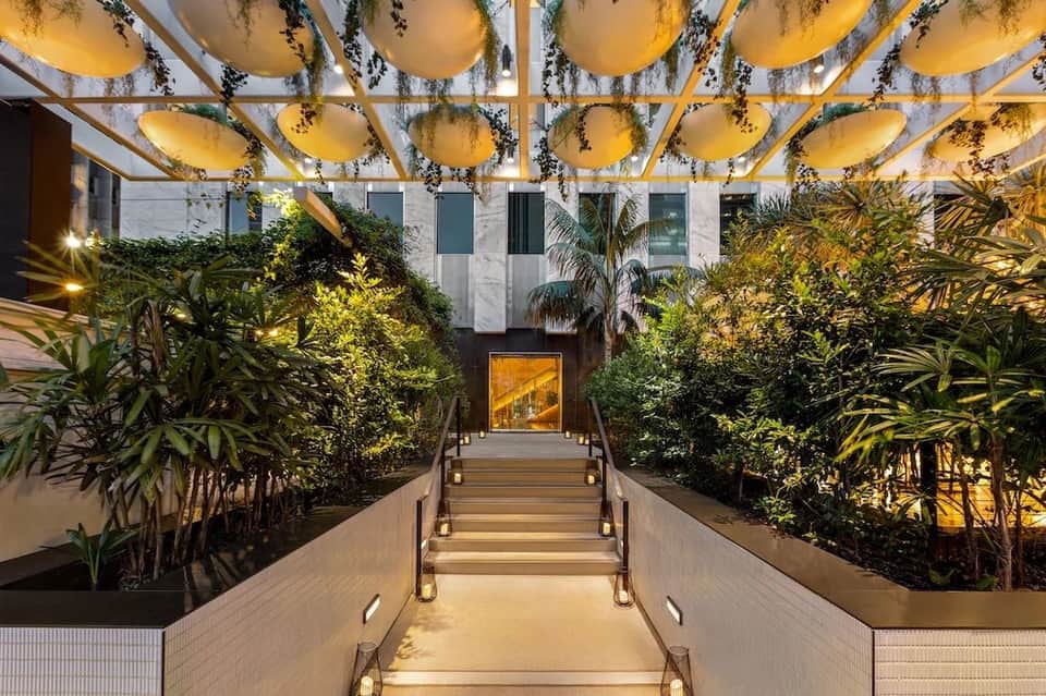 Illuminated covered walkway with lush green plants and hanging golden spheres leading to building entrance