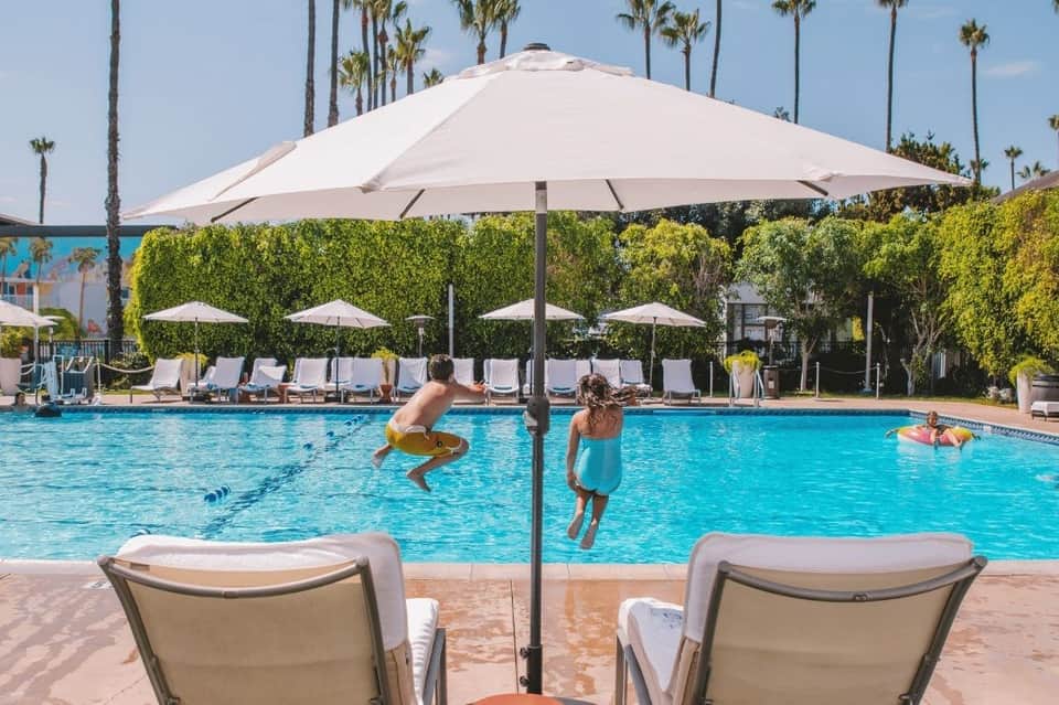 Resort pool with crystal blue water, lounge chairs, large umbrella, and children swimming under palm trees