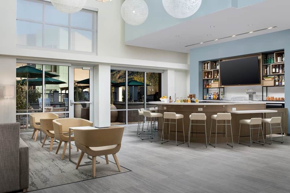 Modern hotel lounge with beige chairs, bar area with shelving, and glass doors opening to outdoor patio