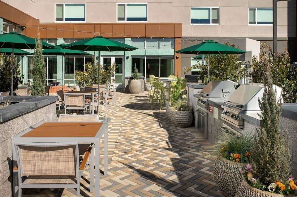 Patio dining area with green umbrellas, outdoor grills, and modern hotel building with herringbone pavers