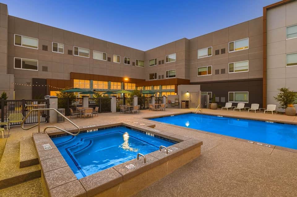 Night view of hotel pool area with spa tub, lounge chairs, modern building facade with lit windows