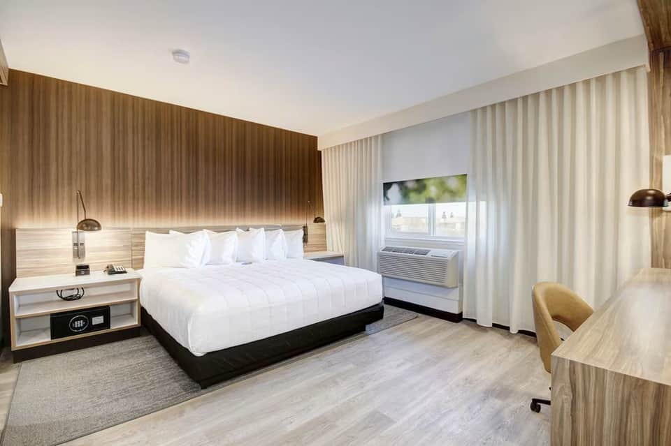 Modern hotel bedroom with queen bed, wooden headboard, light wood flooring, and white curtains framing windows