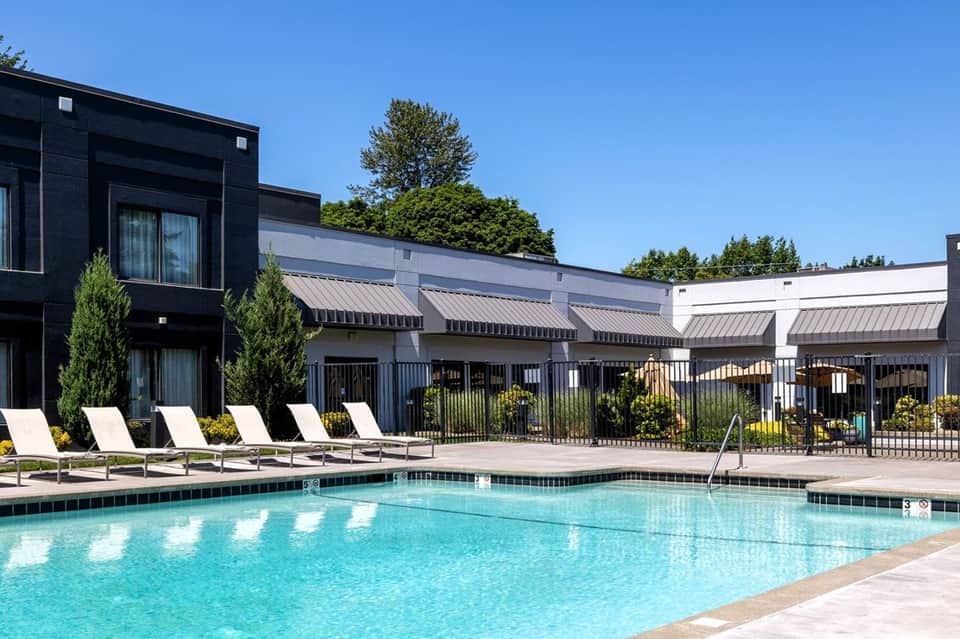 Resort swimming pool with lounge chairs, modern black and white buildings, metal fence, and trees under clear blue sky