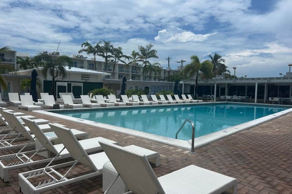 Resort swimming pool with turquoise water, white lounge chairs, palm trees, and multi-story hotel building under blue sky