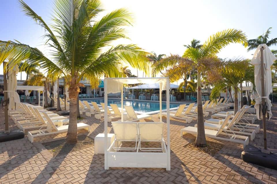 Resort pool with white lounge chairs, palm tree shade structures, pergolas, and calm blue water surrounded by tropical vegetation