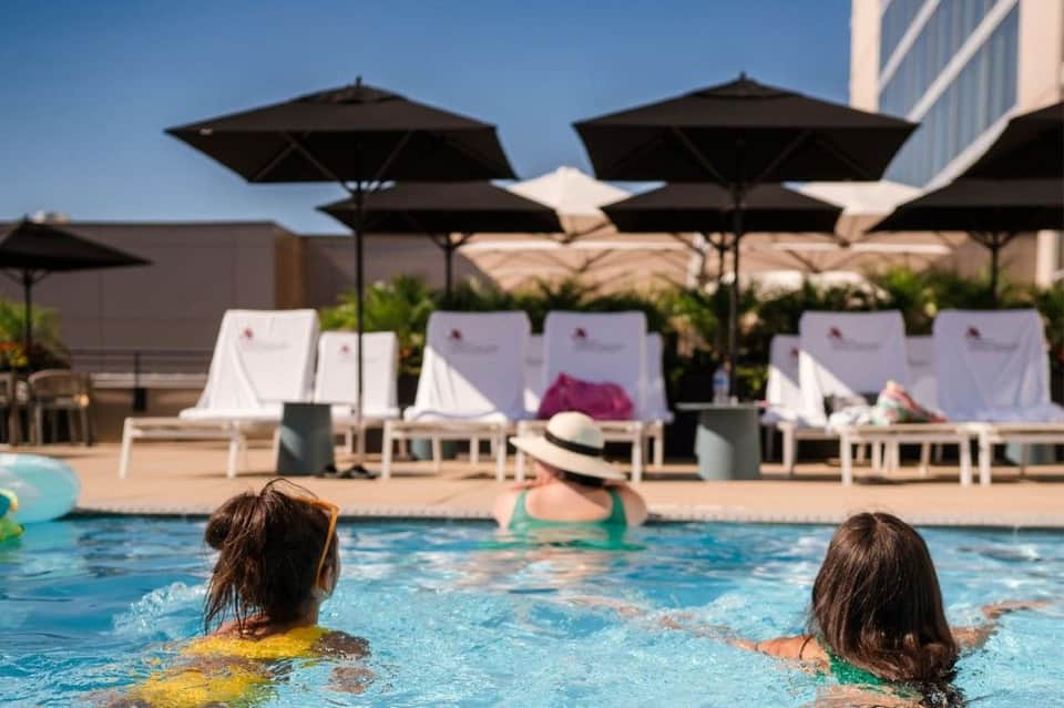 Two children swimming in a bright blue pool with black umbrellas and white lounge chairs visible in the background